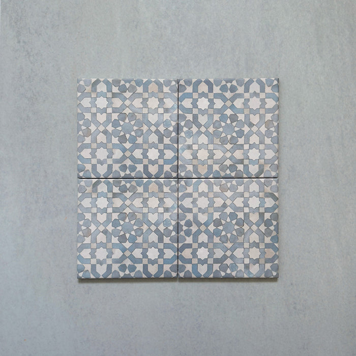Disposición de cuatro azulejos de estilo marroquí Bedouin Tetuan Azul 15x15 cm, mostrando un detallado patrón geométrico de estrellas y cruces en tonos azul, blanco y beige con acabado artesanal y textura sutil sobre fondo claro.