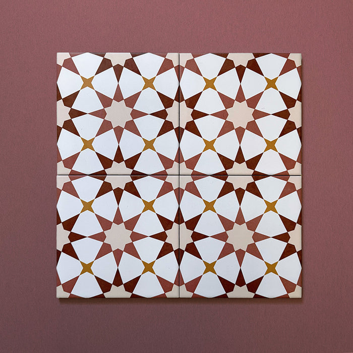 Primer plano de cuatro azulejos cerámicos de estilo marroquí Agraba Cotto 20x20 cm colocados juntos, destacando el patrón geométrico de estrellas en tonos terracota, beige, mostaza y blanco sobre fondo burdeos suave.