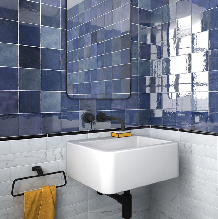 Zona de lavabo en baño con azulejos de pared cerámicos Zellij Azul Cuadrado 13,2 × 13,2 cm de estilo marroquí, mostrando una rica variación en tonos azules con acabado brillante, combinados con lavabo suspendido, grifería negra mate y azulejos inferiores con efecto mármol.