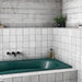 Interior de baño con azulejos de pared cerámicos Zellij Blanco Cuadrado 13,2 × 13,2 cm de estilo marroquí, colocados en un diseño en cuadrícula, con sutil variación tonal y efecto artesanal, combinado con bañera verde, grifería negra y paredes de hormigón.
