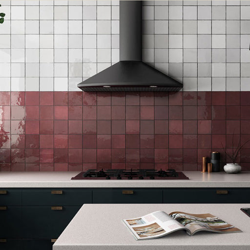 Interior de cocina con azulejos de pared cerámicos Zellij Burdeos Cuadrado 13,2 × 13,2 cm de estilo marroquí, colocados como salpicadero tonal con variación en tonos burdeos intensos y acabado brillante, combinados con azulejos blancos cuadrados en la parte superior, campana extractora negra mate y mobiliario oscuro para crear contraste.