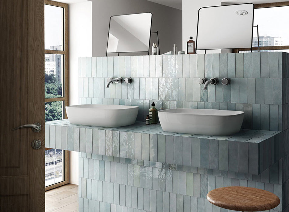 Zona de lavabo en baño con azulejos de pared cerámicos Zellij Aqua Ladrillo 6,5 × 20 cm de estilo marroquí, colocados en disposición vertical en paredes y mueble de lavabo, mostrando una suave variación en tonos azul verdoso, acabado brillante de aspecto artesanal y lavabos dobles modernos con grifería cromada.