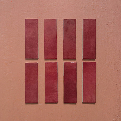 Vista cenital de azulejos de pared cerámicos Zellij Burdeos Ladrillo 6,5 × 20 cm de estilo marroquí, presentados como piezas individuales, destacando los tonos burdeos intensos, la variación natural del color, los bordes ligeramente irregulares y el acabado brillante artesanal sobre un fondo texturizado cálido.