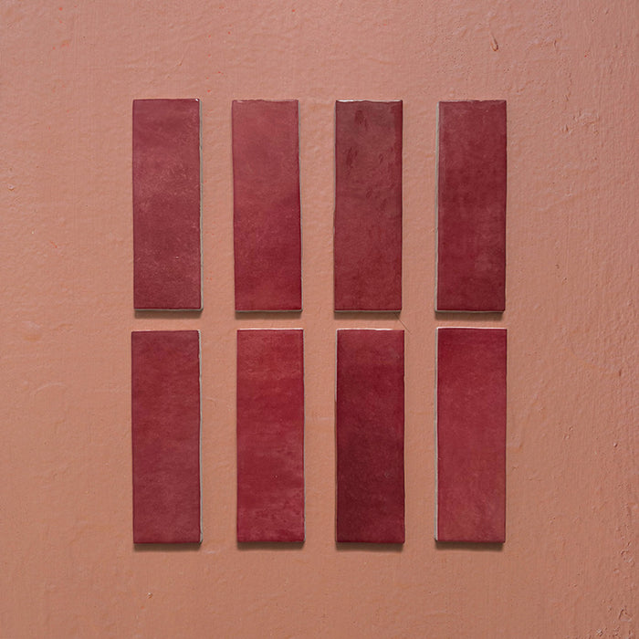 Vista cenital de azulejos de pared cerámicos Zellij Burdeos Ladrillo 6,5 × 20 cm de estilo marroquí, presentados como piezas individuales, destacando los tonos burdeos intensos, la variación natural del color, los bordes ligeramente irregulares y el acabado brillante artesanal sobre un fondo texturizado cálido.