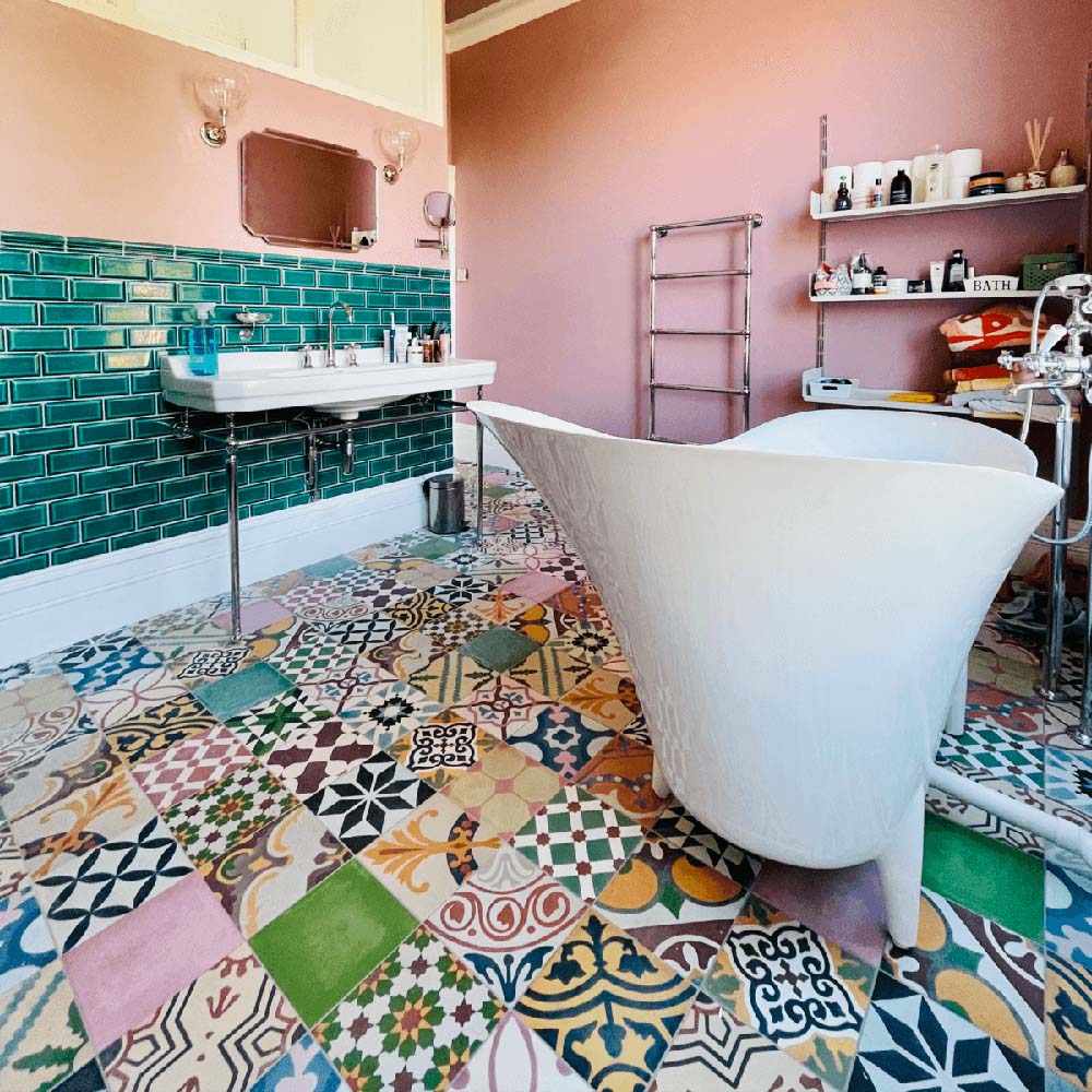Interior de baño con baldosas porcelánicas Fihrdos Patchwork 20x20 cm de estilo marroquí para suelo, mostrando una mezcla de coloridos patrones geométricos, combinadas con azulejos tipo metro verdes en la pared, paredes en rosa empolvado y bañera exenta para crear un suelo protagonista.