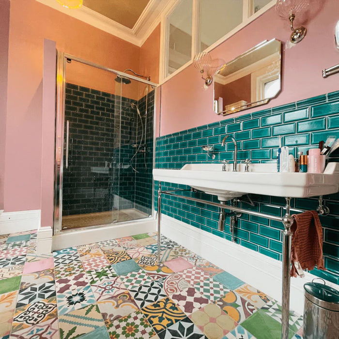 Interior de baño con baldosas porcelánicas Fihrdos Patchwork 20x20 cm de estilo marroquí para suelo, mostrando una mezcla de coloridos patrones geométricos, combinadas con azulejos tipo metro verdes en la pared, paredes en rosa empolvado y bañera exenta para crear un suelo protagonista.