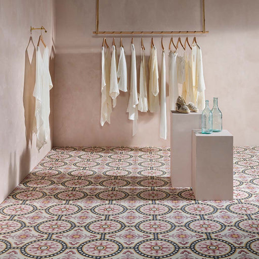 Interior con baldosas de suelo Europa Dortmund 20 × 20 cm de estilo Victoriano Marroquí en tonos rojo, crema, gris y mostaza, combinadas con un perchero con prendas en tonos neutros y botellas decorativas de vidrio en un espacio de estilo boutique.