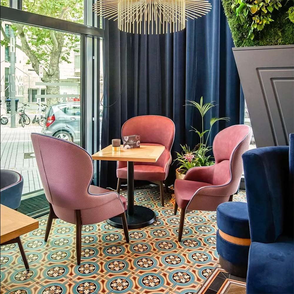 Interior de restaurante con baldosas Europa Helsinki 20 × 20 cm de estilo Victoriano Marroquí en tonos verde azulado, terracota y crema, instaladas como suelo decorativo bajo sillas tapizadas en rosa, combinadas con cortinas azul marino, plantas y mesas de madera natural.