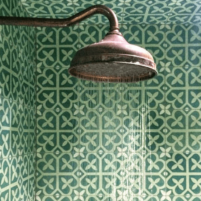 Pared de ducha revestida con baldosas porcelánicas Iris Verde 20x20 cm de estilo Victoriano Marroquí en tonos verdes intensos con patrón geométrico clásico, combinadas con rociador de ducha en latón vintage para un baño decorativo con carácter.