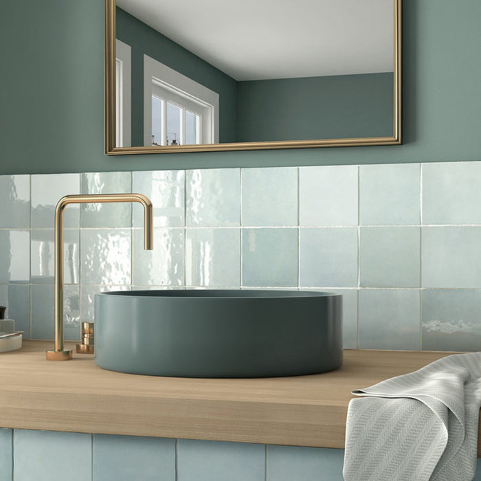 Zona de lavabo en baño con azulejos de pared cerámicos Zellij Aqua Cuadrado 13,2 × 13,2 cm de estilo marroquí, mostrando una suave variación en tonos azules con acabado brillante, combinados con lavabo redondo, grifería de latón, encimera de madera y paredes pintadas en verde.