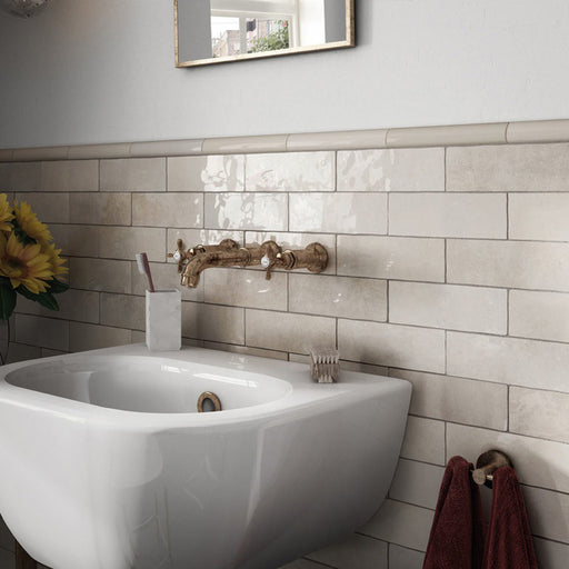 Interior de baño con azulejos de pared cerámicos Zellij Ocre Ladrillo 6,5 × 20 cm de estilo marroquí en tonos neutros cálidos, colocados en un diseño tipo ladrillo con acabado brillante, combinados con grifería de latón empotrada en pared, lavabo blanco y luz natural suave.