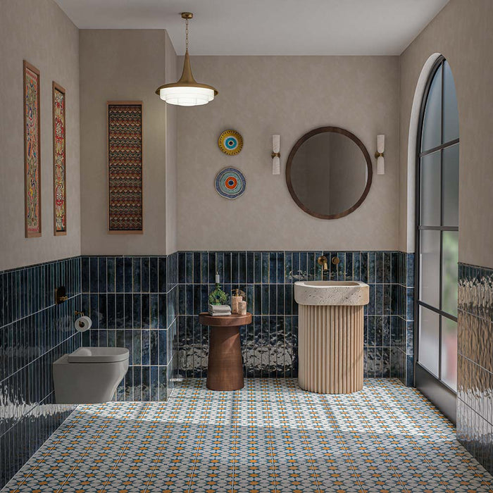Baño lleno de color con azulejos de suelo Agraba Blue and Yellow de 20 × 20 cm de estilo marroquí, con motivos geométricos de estrellas en tonos azul, naranja y crema, combinados con azulejos de pared brillantes en azul intenso, un lavabo acanalado en color crema y grifería en acabado latón.