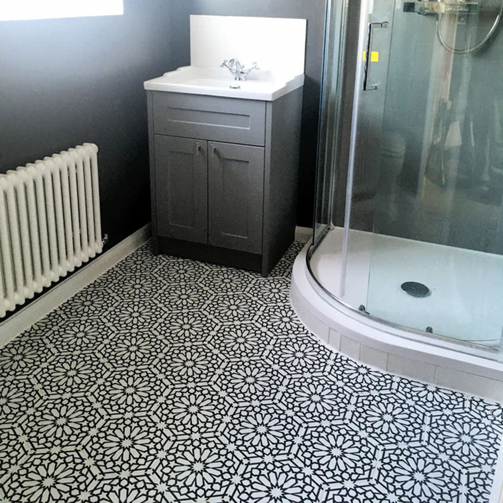 Interior de baño con azulejos cerámicos de estilo marroquí Alhambra Hexagon 18,7x21,6 cm para suelo, mostrando un detallado patrón geométrico en blanco y negro, combinados con mueble gris y mampara de ducha curva de cristal.