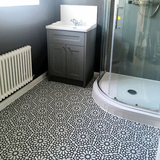 Interior de baño con azulejos cerámicos de estilo marroquí Alhambra Hexagon 18,7x21,6 cm para suelo, mostrando un detallado patrón geométrico en blanco y negro, combinados con mueble gris y mampara de ducha curva de cristal.