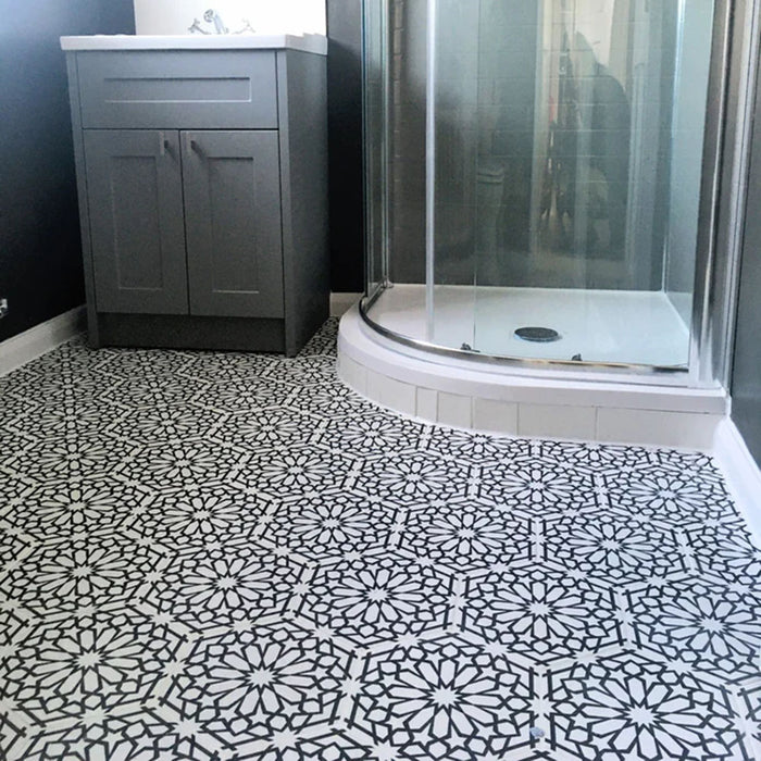Interior de baño con azulejos cerámicos de estilo marroquí Alhambra Hexagon 18,7x21,6 cm para suelo, destacando un patrón geométrico detallado en blanco y negro, combinado con mueble gris y mampara de ducha curva de cristal.