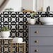 Interior de baño con azulejos cerámicos de estilo marroquí Alhambra Mini Jemia 15x15 cm para pared, mostrando un audaz patrón geométrico en blanco y negro colocado detrás de un mueble gris con detalles en latón y grifería negra mate.