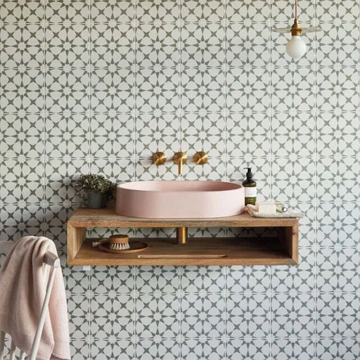 Interior de baño con azulejos cerámicos de estilo marroquí Agraba Sage 20x20 cm para pared, mostrando un suave patrón geométrico en verde salvia colocado detrás de un mueble suspendido con grifería de latón y lavabo en tono rosado.