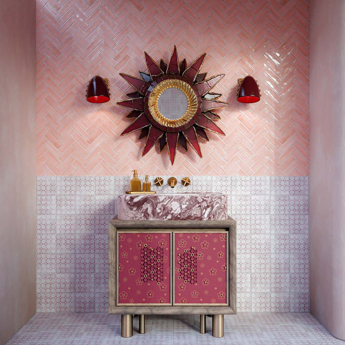 Baño con azulejos Bedouin Mequinez Rose de estilo marroquí para paredes y suelo, combinados con azulejos Atlas Pink Mini Brick Gloss colocados en patrón de espiga. Decorado con un lavabo efecto mármol en tonos rosa, grifería en acabado latón, un espejo decorativo tipo sol y cálidos tonos rosados que crean un espacio atrevido y lleno de personalidad.