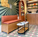 Café setting featuring Geometry Seritina 20x20cm patterned floor tiles with green striped and pink semicircle motifs on a cream base, styled with a wooden bench, terracotta-toned cushion, modern shelving, and a marble-topped coffee table.