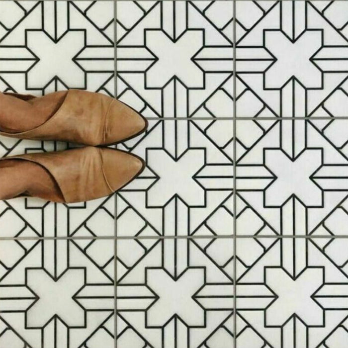 Azulejos de estilo marroquí Alhambra Palacio 20x20 cm con patrón geométrico en cruz en negro sobre base blanca, mostrados desde arriba junto a zapatos en tono camel para destacar el diseño detallado y el acabado mate.