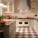 Elegante cocina con azulejos Fizaan Pink Rose de 20 × 20 cm de estilo marroquí en el salpicadero, con delicados motivos geométricos florales en tonos rosa y crema, combinados con muebles en color crema, encimeras efecto mármol burdeos, un suelo de damero en rojo y crema y grifería en acabado latón.