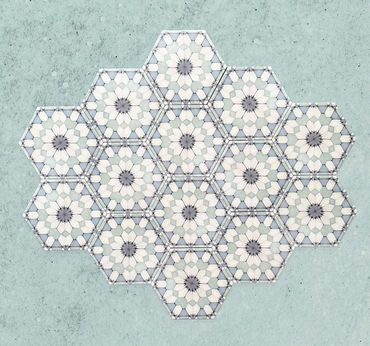 Baldosas hexagonales Maroush Azul Escarcha 18,7x21,6 cm de estilo marroquí dispuestas en grupo, mostrando un patrón floral repetitivo en forma de estrella en azul suave, blanco y gris sobre fondo azul claro texturizado.