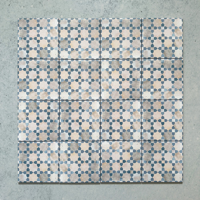 Conjunto de azulejos de estilo marroquí Bedouin Mequinez Azul 15x15 cm para pared y suelo, dispuestos en un patrón geométrico repetido en suaves tonos azul, crema y topo sobre fondo texturizado claro, mostrando el efecto decorativo completo.