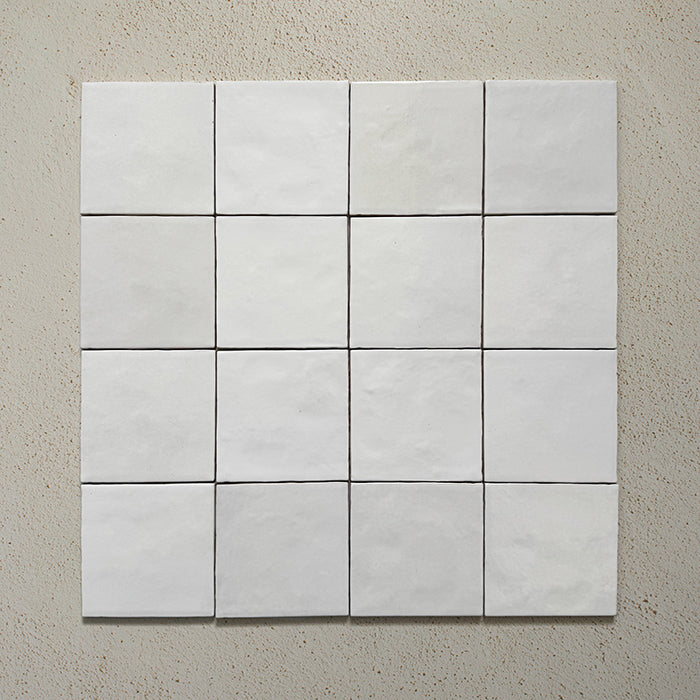 Vista cenital de azulejos de pared cerámicos Zellij Blanco Cuadrado 13,2 × 13,2 cm de estilo marroquí, dispuestos en cuadrícula, mostrando una ligera variación en la superficie, bordes suaves y un acabado brillante con efecto artesanal sobre un fondo neutro texturizado.