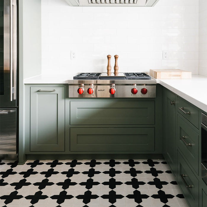 Interior de cocina con azulejos cerámicos de estilo marroquí Alhambra Compass 20x20 cm para suelo, destacando un potente patrón geométrico en blanco y negro, combinado con mobiliario en verde salvia y azulejos tipo metro blancos en la pared.