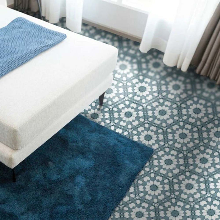 Suelo de dormitorio con baldosas porcelánicas Maroush Azul Arcilla 18,7x21,6 cm en formato hexagonal de estilo marroquí, mostrando un patrón geométrico en azul suave y blanco, combinado con alfombra azul marino y banco tapizado para un acabado decorativo con carácter.