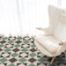 Living room setting featuring Geometry Angles Patterned Tile in 20cm x 20cm, arranged in a repeating geometric pattern with green, black, and off-white triangular shapes. A soft white armchair sits on the tiled floor beside sheer white curtains, creating a bright and airy atmosphere.