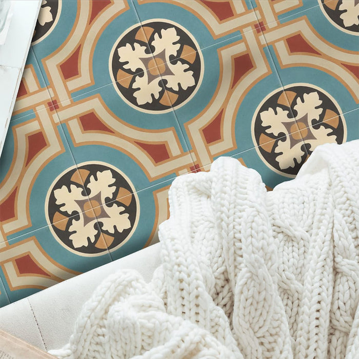 Vista cenital de baldosas de suelo Europa Helsinki 20 × 20 cm de estilo Victoriano Marroquí en tonos verde azulado, terracota, crema y negro, formando un patrón geométrico decorativo, combinadas con una manta de punto suave en un interior acogedor.