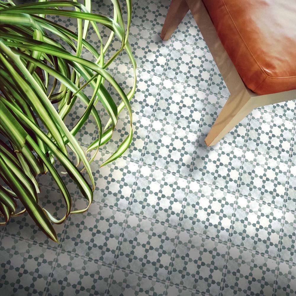 Azulejos decorativos de estilo marroquí Bedouin Mequinez Verde 15x15 cm para suelo, presentados en un ambiente de inspiración con sillón de cuero color camel y planta verde de interior, destacando el patrón geométrico de estrellas en verde suave y tonos neutros con acabado efecto artesanal.