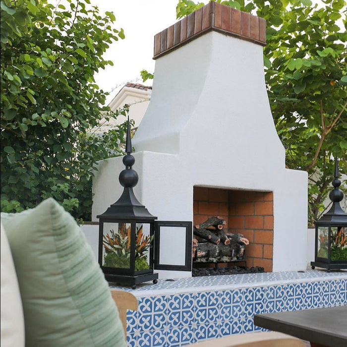 Espacio exterior con chimenea decorada con baldosas porcelánicas Iris Azul 20x20 cm de estilo Victoriano Marroquí, utilizadas como marco decorativo junto a mampostería blanca y vegetación natural.