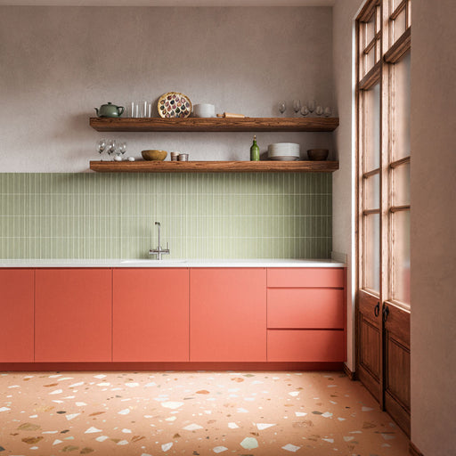 Cocina con azulejos de pared Rippled Brick 3D DECOR Tansy Green Matt de 5 × 20 cm, con una textura acanalada y un suave tono verde, combinados con muebles en tono coral, suelo de estilo terrazo y cálidos detalles en madera.