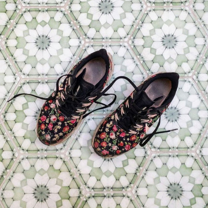 Baldosas Maroush Verde 18,7x21,6 cm de estilo marroquí con patrón floral geométrico en verde suave y blanco, ambientadas con zapatillas estampadas sobre el suelo para un escenario de estilo de vida informal.