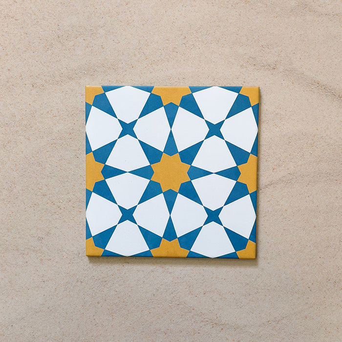 Primer plano del azulejo cerámico de estilo marroquí Agraba Sunshine 20x20 cm, con un llamativo patrón geométrico de estrellas en azul, amarillo mostaza y blanco sobre un fondo arena neutro.