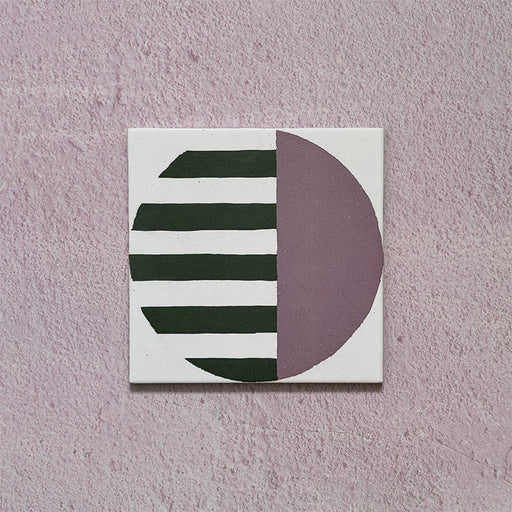 Single tile image showing the Geometry Seritina 20x20cm patterned tile with green horizontal stripes and a mauve semicircle on a cream base, photographed against a textured pink background.