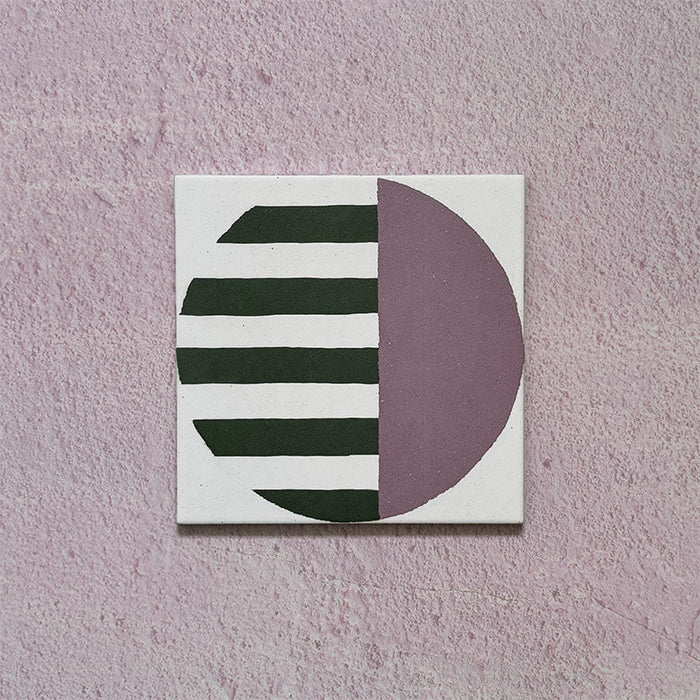Single tile image showing the Geometry Seritina 20x20cm patterned tile with green horizontal stripes and a mauve semicircle on a cream base, photographed against a textured pink background.