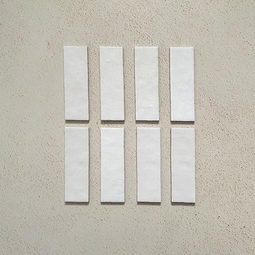 Vista cenital de azulejos de pared cerámicos Zellij Blanco Ladrillo 6,5 × 20 cm de estilo marroquí en color blanco, presentados como piezas individuales con sutil variación tonal y acabado artesanal sobre un fondo neutro texturizado.