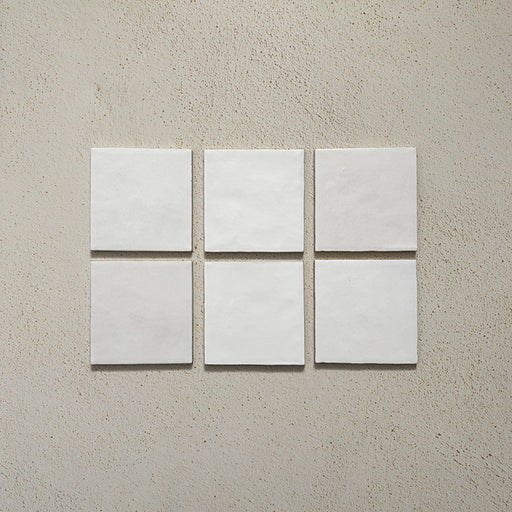 Vista cenital de azulejos de pared cerámicos Zellij Blanco Cuadrado 13,2 × 13,2 cm de estilo marroquí, presentados como piezas individuales, destacando bordes suaves, ligera variación en la superficie y un acabado brillante artesanal sobre un fondo neutro texturizado.