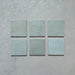 Vista cenital de azulejos de pared cerámicos Zellij Aqua Cuadrado 13,2 × 13,2 cm de estilo marroquí, presentados como piezas individuales, mostrando la suave variación tonal en azul, el acabado brillante artesanal y los bordes sutilmente irregulares para un aspecto auténtico y lleno de carácter.