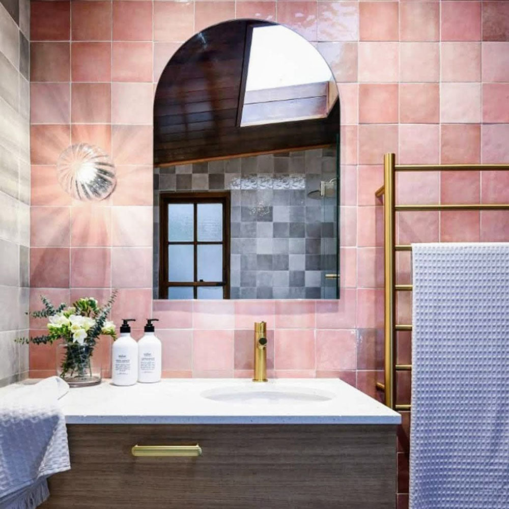 Baño con azulejos de pared cerámicos Zellij Pink Square 13,2 × 13,2 cm de estilo marroquí, mostrando una suave variación tonal y una textura brillante de aspecto artesanal, combinados con grifería de latón, mobiliario de madera cálida y un espejo arqueado.